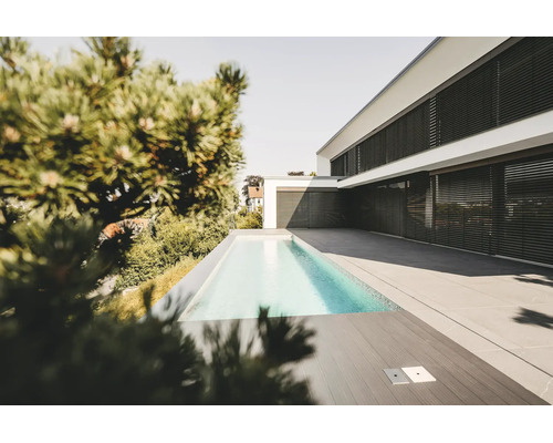 Maison moderne blanche avec stores extérieurs noirs, piscine rectangulaire étroite et vaste terrasse en dalles de pierre grise.