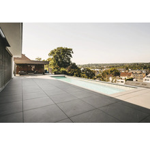Terrasse de toit moderne avec grands carreaux en pierre grise et piscine à débordement. Salon couvert en arrière-plan. Atmosphère lumineuse.