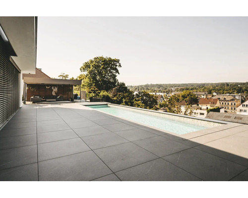 Terrasse de toit moderne avec grands carreaux en pierre grise et piscine à débordement. Salon couvert en arrière-plan. Atmosphère lumineuse.