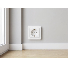 Moderne weiße Schutzkontakt-Steckdose aus Kunststoff an einer Wand über einer weißen Sockelleiste und hellem Holzboden in einem hellen Raum.