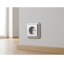 Silberfarbene Schutzkontakt-Steckdose an einer hellen Wand über einer Holzfußleiste montiert.