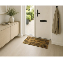 Paillasson Welcome avec motif de feuilles dans une entrée moderne et lumineuse devant une porte d'entrée blanche.