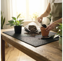 Personne rempotant une plante d'intérieur sur un tapis de jardinage avec du terreau, un pot en terre cuite et des outils sur une table en bois.