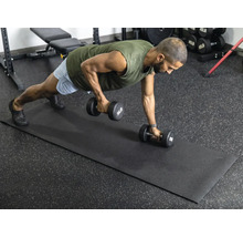 Un homme fait de la musculation avec des haltères sur un tapis de fitness noir dans une salle de sport claire et organisée avec un sol en caoutchouc.