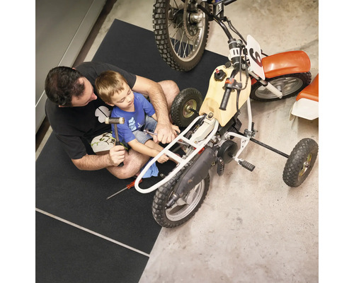 Homme et enfant réparant une moto sur un tapis de protection de sol dans un atelier.