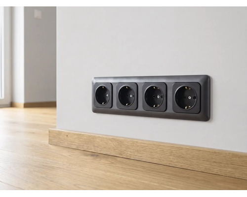 Schwarze 4-fach Schutzkontakt-Steckdose an einer hellen Wand über einer Fußleiste aus Holz in einem hellen, modern gestalteten Raum.