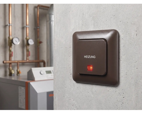 Interrupteur de secours pour chauffage brun avec inscription HEIZUNG et témoin lumineux rouge sur un mur en béton dans une chaufferie.