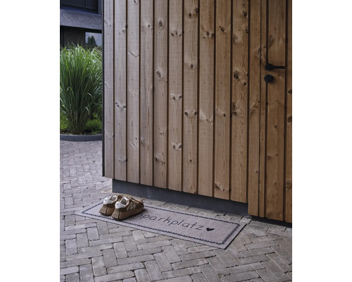 Paillasson avec inscription Schuhparkplatz et un cœur devant une façade en bois sur un sol pavé avec des chaussures.