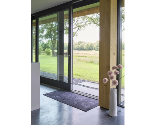 Paillasson rectangulaire à motif de feuilles devant une porte coulissante en verre moderne avec vue sur le jardin et un vase décoratif.