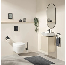 Salle de bain moderne avec WC suspendu, vasque à poser blanche sur meuble sous-vasque beige, robinetterie noire, miroir ovale et tapis gris.