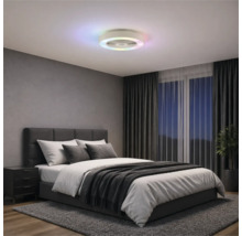 Ventilateur de plafond moderne avec anneau lumineux à LED coloré dans une chambre avec lit rembourré gris, rideaux sombres et tapis.