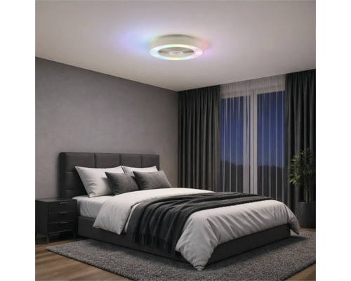 Ventilateur de plafond moderne avec anneau lumineux à LED coloré dans une chambre avec lit rembourré gris, rideaux sombres et tapis.