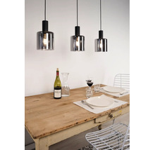 Trois suspensions avec des abat-jour cylindriques en verre fumé suspendues au-dessus d'une table en bois dressée dans une salle à manger lumineuse.