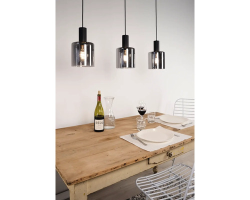 Trois suspensions avec des abat-jour cylindriques en verre fumé suspendues au-dessus d'une table en bois dressée dans une salle à manger lumineuse.