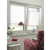 Weißes Plissee an einem zweiflügeligen Fenster in einem hellen Zimmer mit rotem Sessel, Tischchen und Blumen.