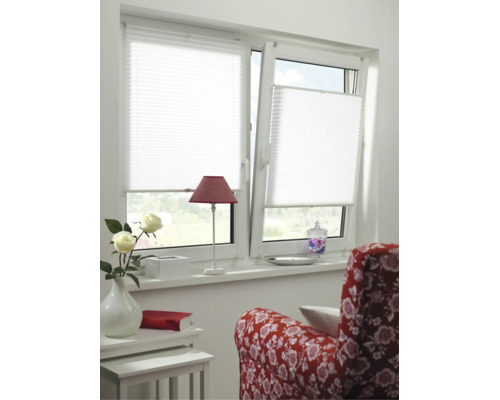 Weißes Plissee an einem zweiflügeligen Fenster in einem hellen Zimmer mit rotem Sessel, Tischchen und Blumen.