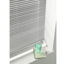 Jalousie mit horizontalen Lamellen vor einem hellen Fenster, die Schatten auf eine Fensterbank mit Glas und Notizblock wirft.