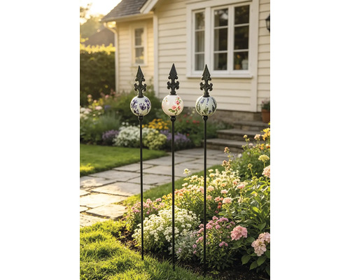 Trois tuteurs de jardin décoratifs en métal avec boules en céramique décorées et pointes en fleur de lys dans un parterre fleuri.