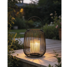 Lanterne noire en fil métallique avec insert en verre et bougie allumée sur une terrasse en bois dans un jardin.