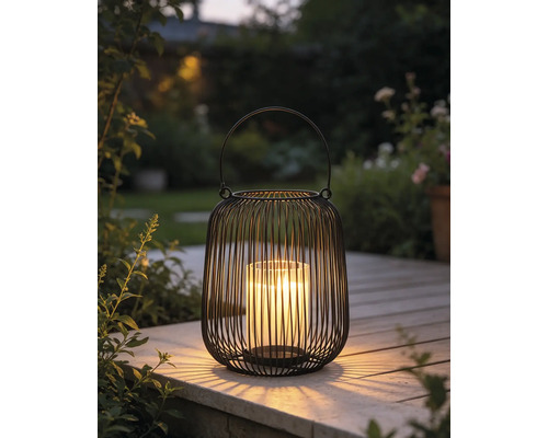 Lanterne noire en fil métallique avec insert en verre et bougie allumée sur une terrasse en bois dans un jardin.
