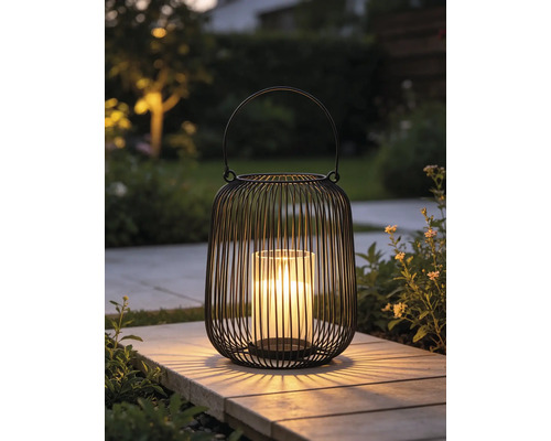 Lanterne moderne en fil métallique noir avec insert en verre et bougie allumée sur une dalle de jardin.