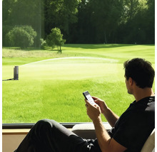 Homme contrôlant un arroseur de jardin intelligent via smartphone en regardant une grande pelouse verte.