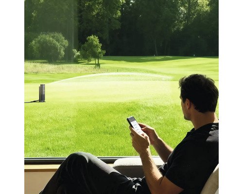 Homme contrôlant un arroseur de jardin intelligent via smartphone en regardant une grande pelouse verte.