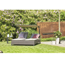 Bain de soleil double en résine tressée avec pare-soleil et coussins dans un jardin lumineux avec pelouse, arbustes en fleurs et fontaine murale.