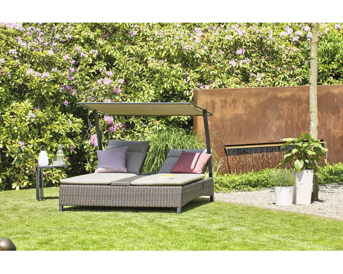 Bain de soleil double en résine tressée avec pare-soleil et coussins dans un jardin lumineux avec pelouse, arbustes en fleurs et fontaine murale.