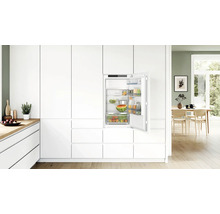 Réfrigérateur encastrable intégré dans une cuisine moderne blanche avec porte ouverte, étagères en verre remplies et tiroir à légumes.