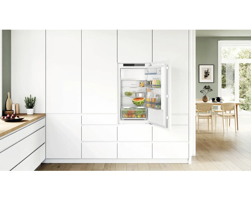 Réfrigérateur encastrable intégré dans une cuisine moderne blanche avec porte ouverte, étagères en verre remplies et tiroir à légumes.