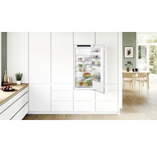 Réfrigérateur encastrable dans une cuisine blanche moderne avec porte ouverte, rempli d aliments, logo Bosch visible à l intérieur.