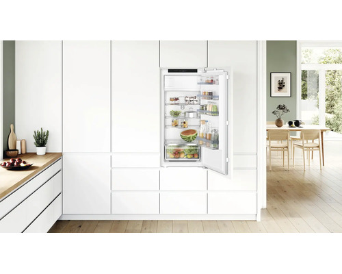 Réfrigérateur encastrable dans une cuisine blanche moderne avec porte ouverte, rempli d aliments, logo Bosch visible à l intérieur.