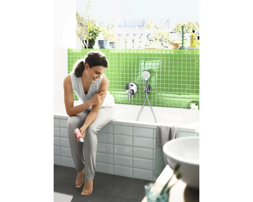 Femme assise sur le bord d'une baignoire dans une salle de bains moderne avec des mosaïques vertes et un mitigeur de baignoire avec douchette.