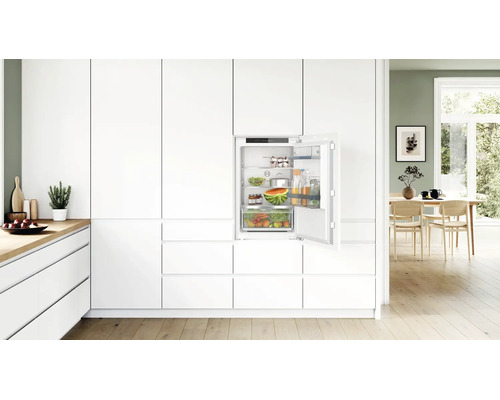 Réfrigérateur encastrable dans une cuisine moderne blanche avec porte ouverte et étagères remplies.