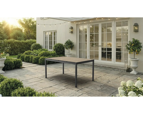 Table de jardin avec plateau en bois et cadre en métal sur une terrasse pavée devant une maison avec de grandes portes-fenêtres.