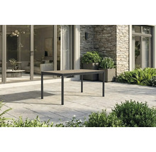 Table de jardin rectangulaire moderne avec cadre en métal et plateau aspect bois sur une terrasse en pierre devant une maison.