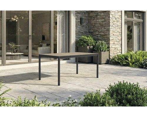 Table de jardin rectangulaire moderne avec cadre en métal et plateau aspect bois sur une terrasse en pierre devant une maison.