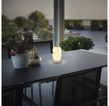 Eine moderne LED Tischleuchte mit Tragegriff steht auf einem Gartentisch auf einer Terrasse mit Pflanzen im Hintergrund.