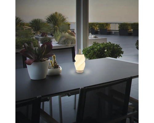 Eine moderne LED Tischleuchte mit Tragegriff steht auf einem Gartentisch auf einer Terrasse mit Pflanzen im Hintergrund.