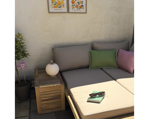 Gemütliche Loungeecke im Freien mit Holzsofa, Beistelltisch, Deko-Kissen, einer kleinen Lampe und Zitrusbildern an der strukturierten Wand.