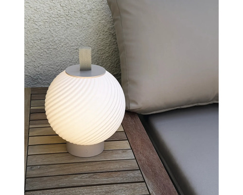 Moderne runde LED Tischleuchte fuer den Aussenbereich mit Trageschlaufe auf einem Holztisch neben einem Polster.