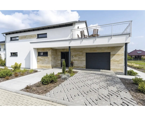 Maison individuelle moderne avec revêtement en pierre naturelle, garage, balcon et pavage en dalles gazon dans le jardin devant.