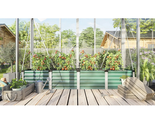 Un potager surélevé en métal dans une serre, planté de tomates, entouré d'outils de jardinage et de caisses en bois sur un sol en bois.