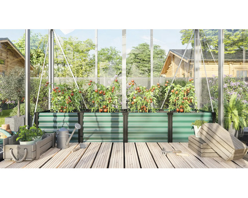 Grand bac à plantes surélevé en métal dans une serre avec des plants de tomates sur une terrasse en bois avec arrosoir et caisses en bois.