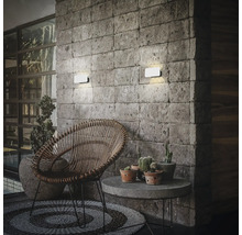Zwei moderne LED Außenwandleuchten montiert an einer Steinwand über einem gemütlichen Lounge Bereich mit Rattansessel und Beistelltisch.