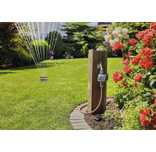 Garten bewässern mit Gardena Bewässerungssteuerung an einem Holzpfosten in einem Garten mit Rasensprenger und blühenden Rosen.