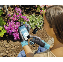Eine Person trägt Gartenhandschuhe und bedient ein Smartphone mit der Gardena Smart System App in einem Garten mit blühenden Pflanzen.