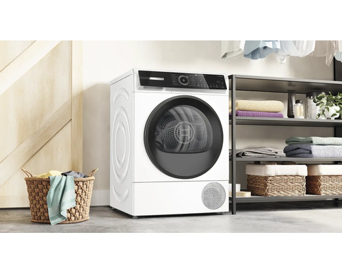 Un sèche-linge Bosch est installé dans une buanderie moderne avec des étagères, des paniers remplis et des vêtements en arrière-plan.