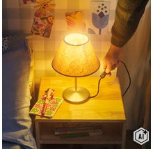 Eine Nachttischlampe mit Textilschirm steht auf einem Holznachttisch und wird von einer Hand eingeschaltet. (Image AI modified or generated)